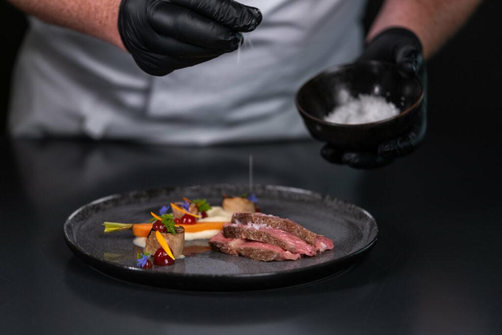 At STURM, sustainable cuisine is served. The chef, wearing gloves, salts a plate of organic meat.