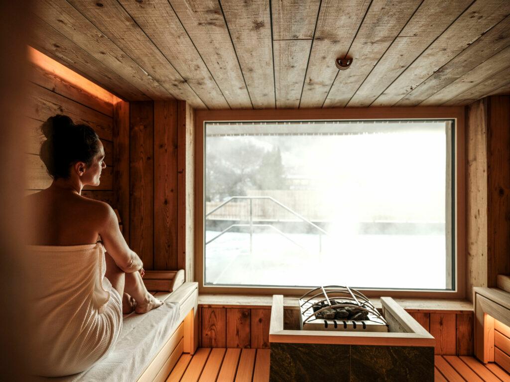 A woman sits in the sauna and looks out at the snow-covered landscape. The sauna is powered by solar and hydroelectric energy.