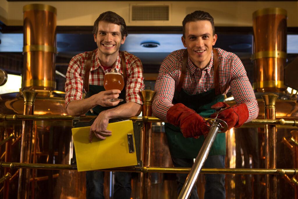 Workers of beer factory posing near bronzed equipment, kettles and pipes. Handsome brewers wearing in checked shirts and aprons looking at camera and smiling. Men holding beer glass and folder.