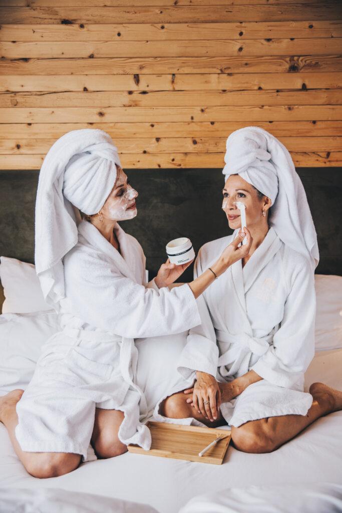 Friends enjoying a glow-up in a hotel sit in robes and try on face masks.