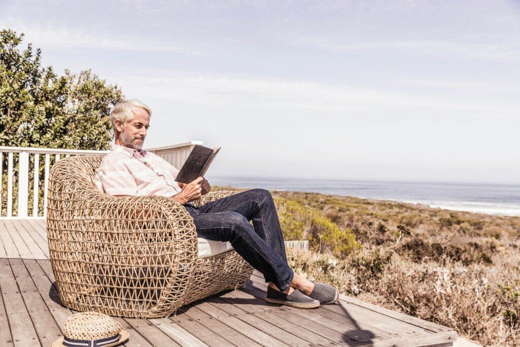 A man sits in a wicker chair reading a book while overlooking the beach