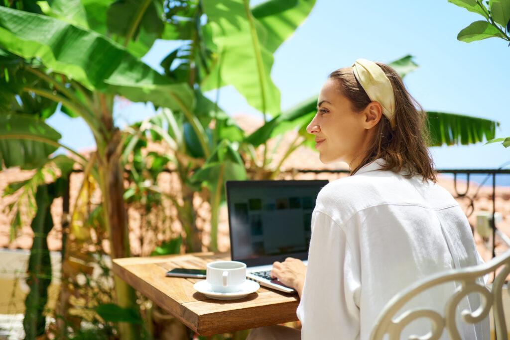 Junge schöne Frau sitzt auf der Terrasse am Morgen im Hotel, mit Laptop-Computer, während einer nachhaltigen Workation unter Palmen