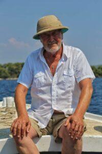 An elderly man in a hat and white shirt sits on a boat under a sunny blue sky. His expression is relaxed, with the sea and trees in the background.