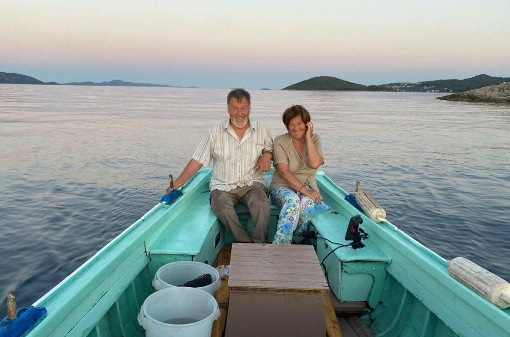 A couple sits smiling in a turquoise boat on calm water during sunset. The horizon is dotted with distant islands, creating a serene, tranquil scene.