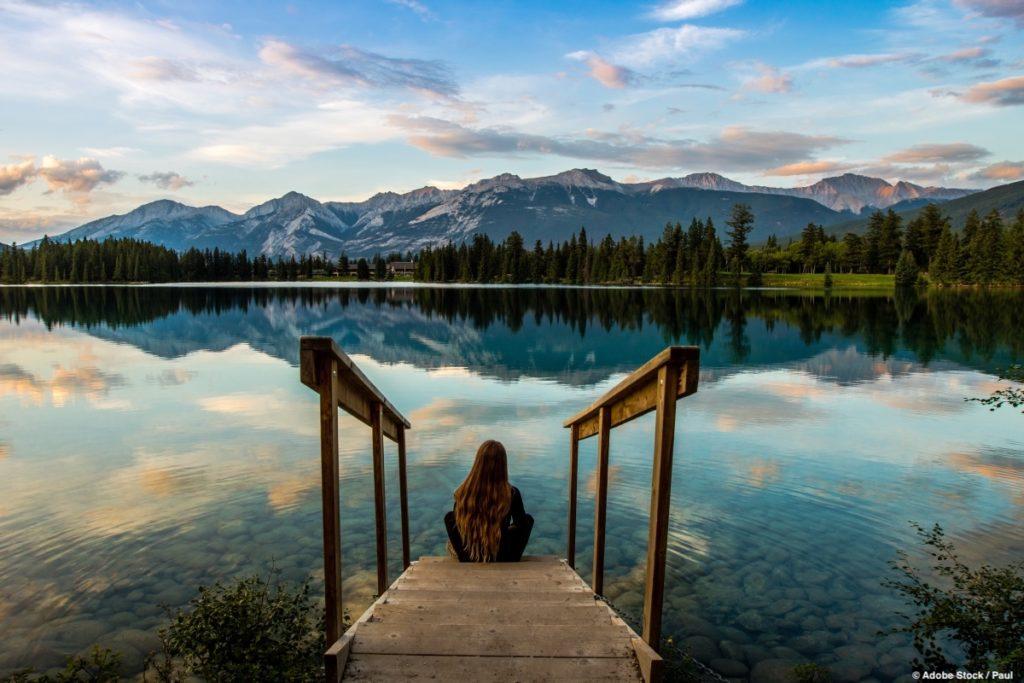 A woman sits on a wooden pier and looks out over a calm lake.Sustainable travel