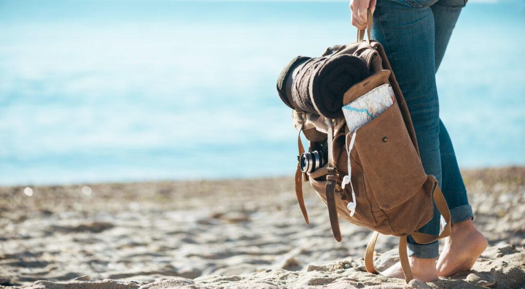 You can see the feet of a traveler standing on the beach. The sea is visible in the background. She holds a small backpack with a rolled up blanket in her hand.