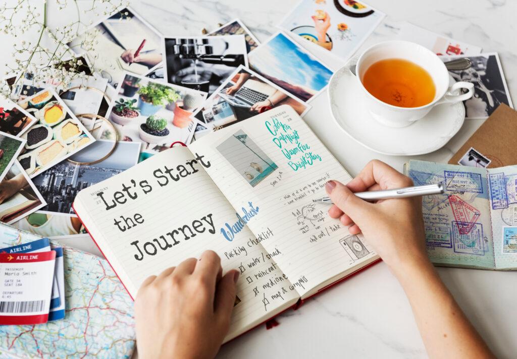 On a table covered with a large map and photos lies a notebook on which is written "Let's start the journey". Two hands are in the picture, one holding a pen. There is also a cup of tea on the table. | We give tips for sustainable travel.