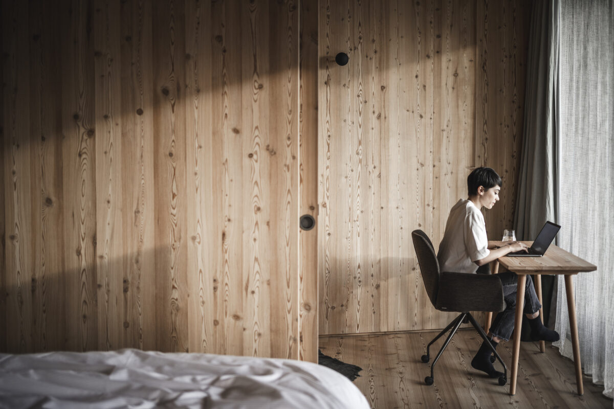 A person is sitting at a wooden desk in a minimalist room with light wood walls, concentrating on a laptop. Soft light streams through a large window, creating a calm atmosphere.