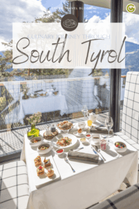 Elegant table setting at the sustainable Hotel my Arbor near Brixen – with diverse dishes and drinks, typical for South Tyrol, on a white tablecloth, overlooking a scenic mountain view under a bright sky. Cozy, inviting atmosphere.