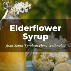 Glass bottle and jar with elderflower syrup, garnished with elderflowers and lemon slice, set against a blurred green background. Text: “Elderflower Syrup from South Tyrolean Hotel Weihrerhof.”