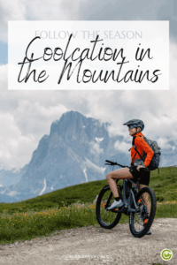 A person in cycling gear and helmet stands with a mountain bike on a grassy path. Snow-capped mountains of the Dolomites tower under cloudy skies, conveying adventure. Text overlay reads "Coolcation in the Mountains."