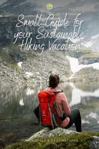 A person with a red backpack sits by a serene mountain lake, surrounded by green slopes and patches of snow, promoting a sustainable hiking vacation.